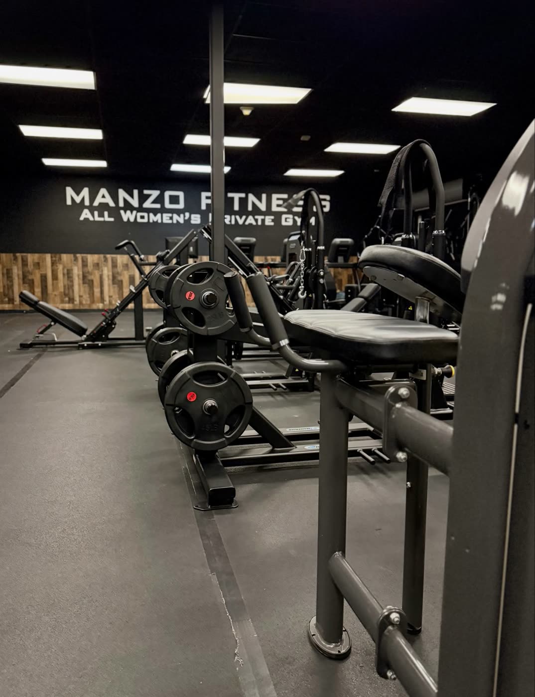 Plate-loaded strength machines in front of the Manzo Fitness wall sign